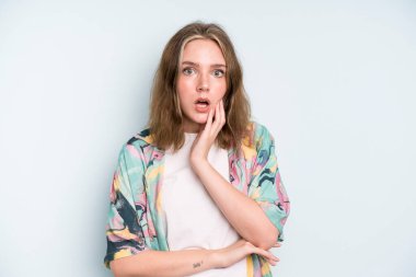 caucasian pretty woman open-mouthed in shock and disbelief, with hand on cheek and arm crossed, feeling stupefied and amazed