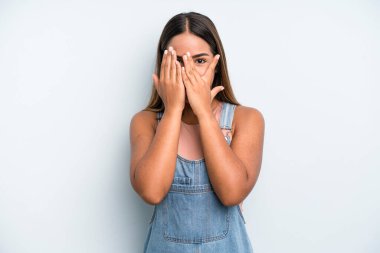 hispanic latin pretty woman covering face with hands, peeking between fingers with surprised expression and looking to the side