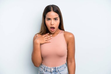 hispanic latin pretty woman feeling shocked, astonished and surprised, with hand on chest and open mouth, saying who, me?