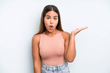 hispanic latin pretty woman looking surprised and shocked, with jaw dropped holding an object with an open hand on the side