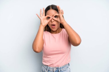 hispanic latin pretty woman feeling shocked, amazed and surprised, holding glasses with astonished, disbelieving look