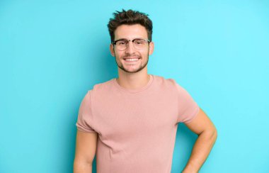 Genç yakışıklı adam mutlu bir şekilde gülümsüyor, eli kalçasında ve kendinden emin, olumlu, gururlu ve arkadaş canlısı.