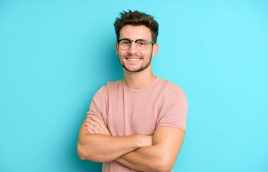 Genç yakışıklı adam kameraya çapraz kollarıyla gülümsüyor ve mutlu, kendinden emin, tatmin olmuş bir ifade, yan bakış.