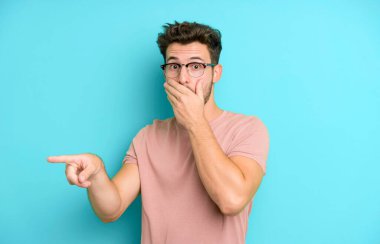 Genç yakışıklı adam kendini mutlu, şaşkın ve şaşırmış hissediyor. Ağzını eliyle kapatıyor ve yanal kopya alanını işaret ediyor.