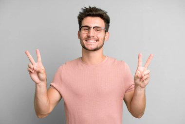 Genç yakışıklı adam gülümsüyor ve mutlu görünüyor, arkadaş canlısı ve memnun, işaret eden zafer ya da barış iki elle