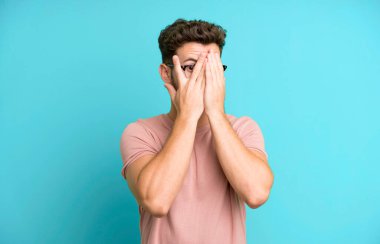 Genç yakışıklı adam elleriyle yüzünü kapatıyor, parmaklarının arasından şaşkın bir ifadeyle yan tarafa bakıyor.