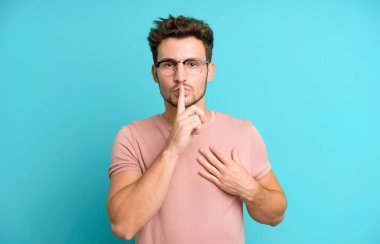 Genç ve yakışıklı adam ciddi görünüyor ve parmaklarını dudaklarına bastırıp sessizlik ya da sessizlik talep ediyor, sır saklıyor.