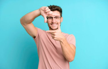 Genç yakışıklı adam mutlu, arkadaş canlısı ve pozitif hissediyor, gülümsüyor ve elleriyle bir portre veya fotoğraf çerçevesi yapıyor.