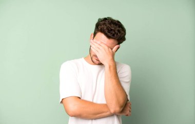 Genç ve yakışıklı bir adam stresli, utanmış ya da üzgün görünüyor, baş ağrısı var, yüzünü eliyle kapatıyor.