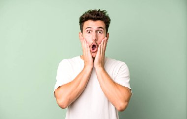 Genç yakışıklı adam şok olmuş ve korkmuş hissediyor, ağzı açık ve elleri yanaklarında korkmuş görünüyor.