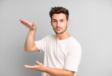 Genç yakışıklı bir adam iki eli de yan tarafında fotokopi alanı olan bir nesneyi tutuyor, bir nesneyi gösteriyor, sunuyor ya da reklamını yapıyor.