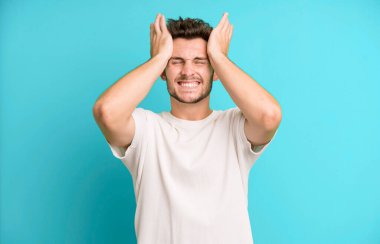 Genç ve yakışıklı bir adam stresli ve endişeli, depresyonda ve hayal kırıklığına uğramış, iki elini de başa kaldırıyor.