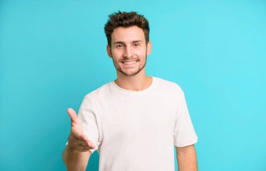 Genç yakışıklı adam gülümsüyor, mutlu görünüyor, kendinden emin ve arkadaş canlısı, bir anlaşmaya varmak için el sıkışıyor, işbirliği yapıyor.