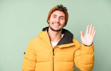 Yakışıklı genç mutlu gülümsüyor, el sallıyor, sizi selamlıyor ve selamlıyor. Kış giysileri ve soğuk konsept
