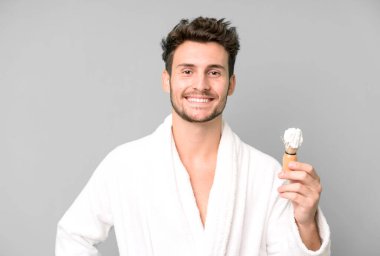 Yakışıklı genç mutlu bir şekilde gülümsüyor, eli kalçasında ve kendine güvenen bornoz giyiyor ve tıraş konsepti kullanıyor.