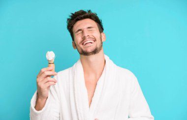 Yakışıklı genç, komik bir şakaya yüksek sesle gülüyor bornoz giyiyor ve traş konsepti kullanıyor.