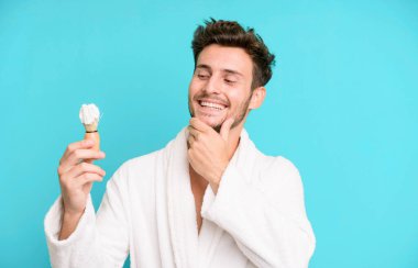 Mutlu, kendine güvenen, elleri çenesinde, bornoz giymiş ve traş konseptiyle gülümseyen yakışıklı bir genç.