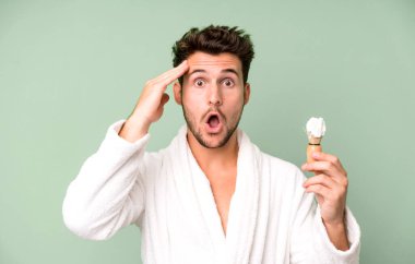 Yakışıklı genç mutlu görünüyor, şaşırmış ve şaşırmış bornoz giyiyor ve traş oluyor.