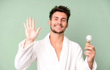 Yakışıklı genç, mutlu bir şekilde gülümsüyor, el sallıyor, seni selamlıyor ve bornoz giyip traş oluyor.