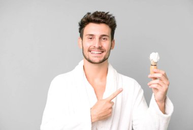 Yakışıklı genç neşeyle gülümsüyor, mutlu hissediyor ve bornoz giyip tıraş konseptini işaret ediyor.