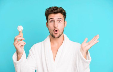 Yakışıklı genç şaşkın ve şaşkın görünüyor, çenesi düşmüş bornoz ve traş konsepti giymiş bir nesneyi tutuyor.