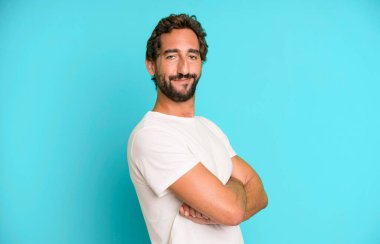 Genç çılgın adam kameraya çapraz kollarla gülümsüyor ve mutlu, kendinden emin, tatmin olmuş bir ifade, yan bakış.