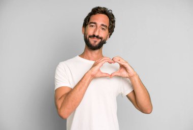 Genç çılgın adam gülümsüyor ve mutlu, sevimli, romantik ve aşık, iki eliyle kalbi şekillendiriyor.
