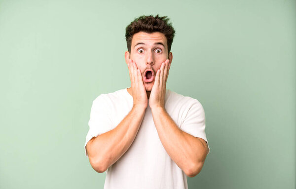 young handsome man feeling shocked and scared, looking terrified with open mouth and hands on cheeks