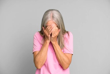 Gri saçlı, yaşlı, üzgün, sinirli ve depresif bir kadın. İki eliyle yüzünü kapatıyor, ağlıyor.