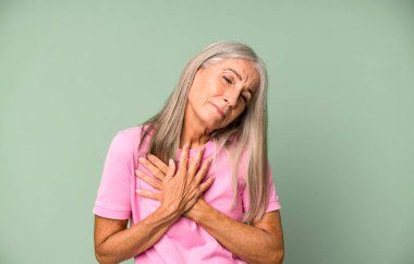 Gri saçlı, üzgün, kalbi kırık, iki elini de kalbe yakın tutan, ağlayan ve depresyonda hisseden yaşlı bir kadın.