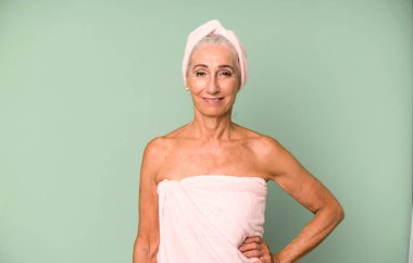 Gri saçlı güzel kadın mutlu bir şekilde gülümsüyor, eli kalçasında ve kendinden emin. Bornoz ve havlu giyiyor. güzellik konsepti