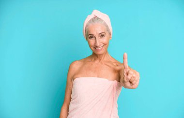 Gri saçlı güzel kadın gülümsüyor ve arkadaş canlısı görünüyor, bir numarayı gösteriyor. Bornoz ve havlu giyiyor. güzellik konsepti