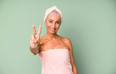 Gri saçlı güzel kadın gülümsüyor ve arkadaş canlısı görünüyor, iki numarayı gösteriyor. Bornoz ve havlu giyiyor. güzellik konsepti