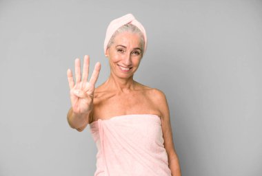 Gri saçlı güzel kadın gülümsüyor ve arkadaş canlısı görünüyor, dört numarayı gösteriyor. Bornoz ve havlu giyiyor. güzellik konsepti