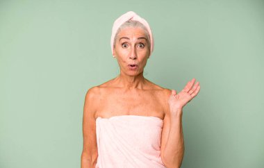 Gri saçlı güzel kadın şaşırmış ve şok olmuş görünüyor. Çenesi düşmüş ve elinde bir nesne var. Bornoz ve havlu giyiyor. güzellik konsepti