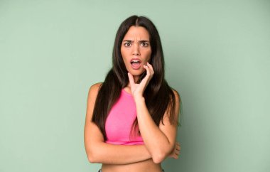İspanyol güzel kadın şok ve inançsızlık içinde ağzı açık, elleri yanağında ve kolları çaprazlanmış, sersemlemiş ve şaşırmış hissediyor