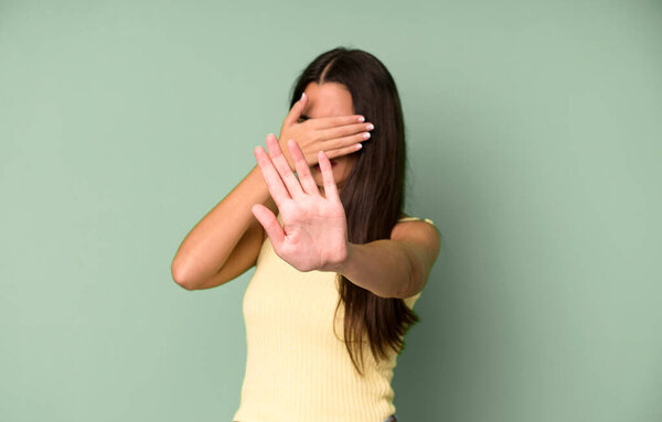 hispanic pretty woman covering face with hand and putting other hand up front to stop camera, refusing photos or pictures