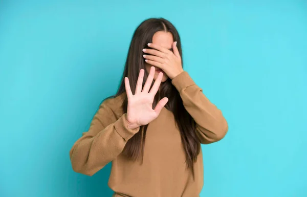 hispanic pretty woman covering face with hand and putting other hand up front to stop camera, refusing photos or pictures