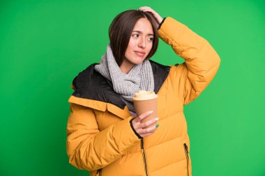 Genç, güzel bir kadın şaşkın ve kafası karışmış hissediyor. Kafasını kaşıyor, palto giyiyor ve elinde kahve tutuyor.