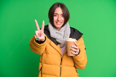 Genç ve güzel bir kadın gülümsüyor ve mutlu görünüyor. Zafer veya huzur simgesi. Ceket giyiyor ve elinde kahve tutuyor.