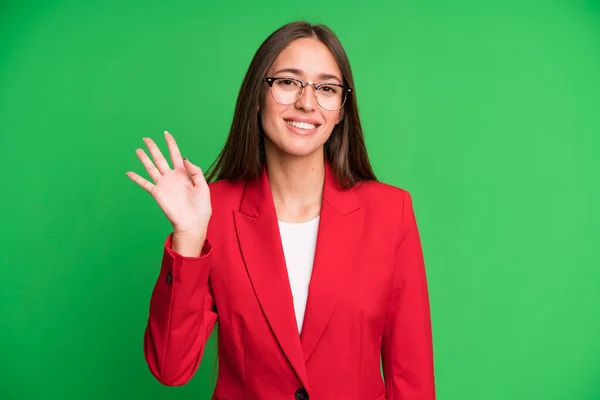 young pretty woman smiling happily, waving hand, welcoming and greeting you wearing a blazer. business concept