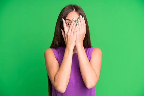 hispanic pretty woman covering face with hands, peeking between fingers with surprised expression and looking to the side