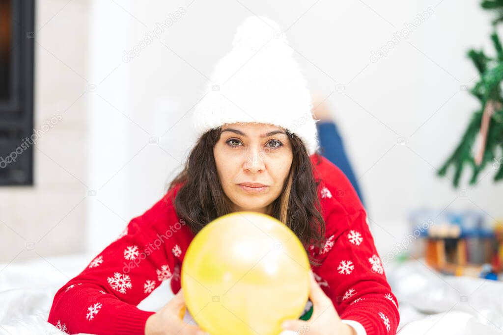 Beautiful woman in her forties wearing white hat and holding yellow balloon in front of a Christmas tree