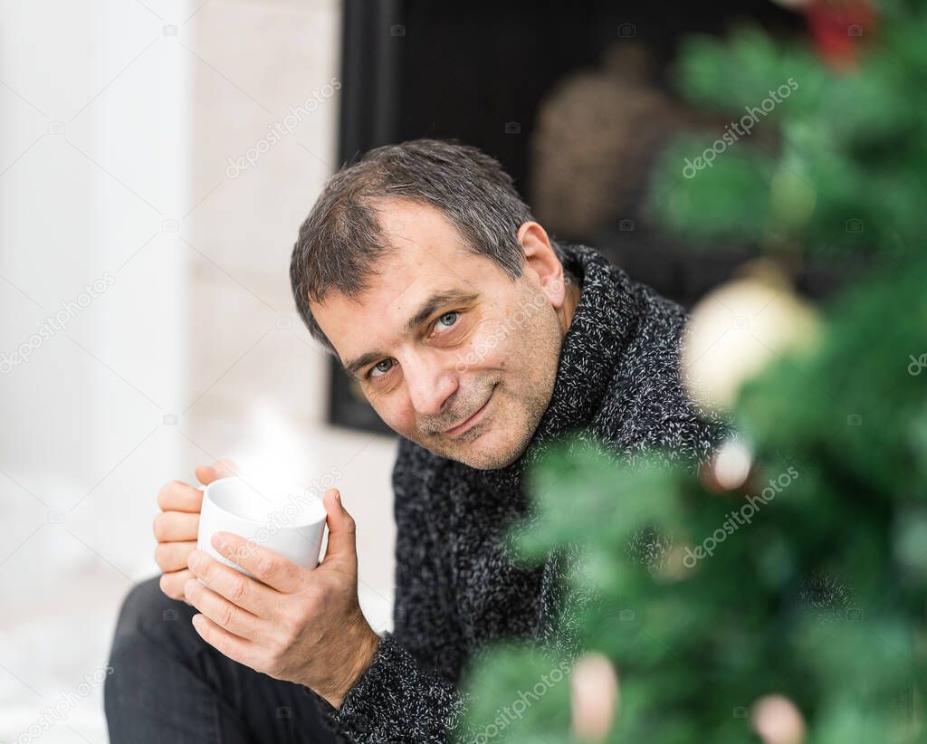 Male in his fifties holding a cup with hot beverage behind a Christmas tree.