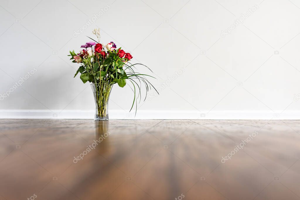 A vase with a rose bouquet placed on hardwood floor next to a wall with baseboard in an empty domestic room.