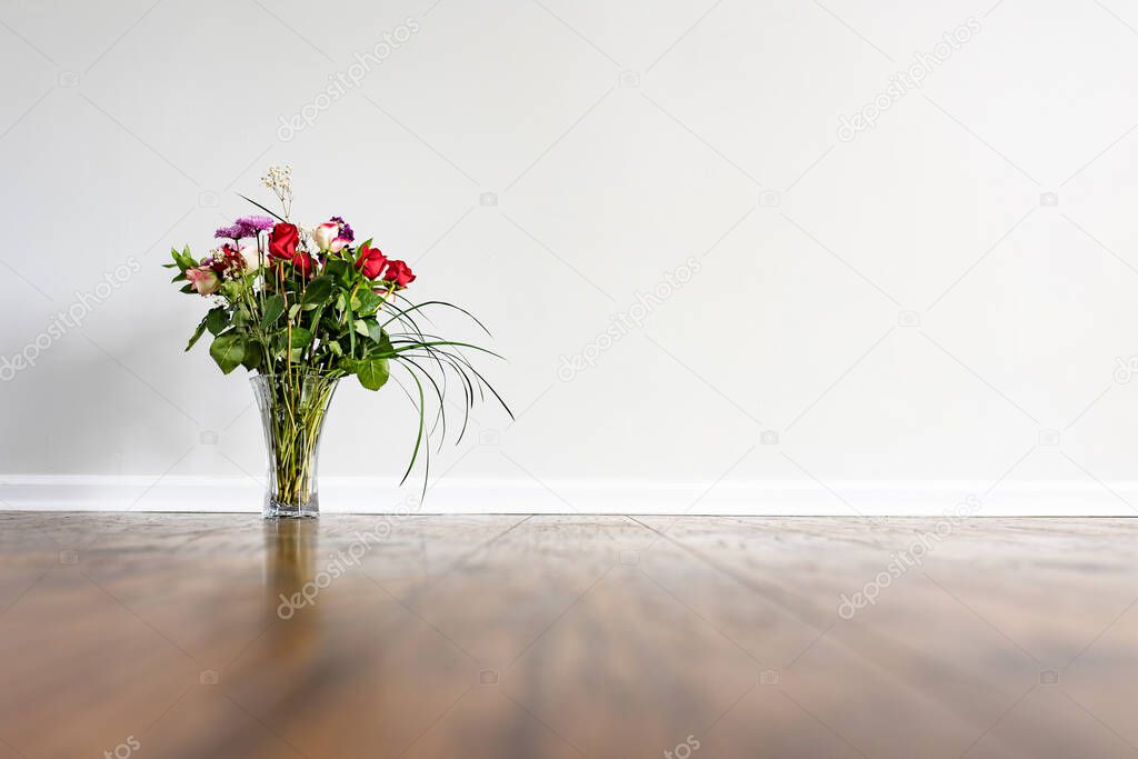 A bouquet with roses and decorative long grass leaves placed on the hardwood floor of empty domestic room with white wall