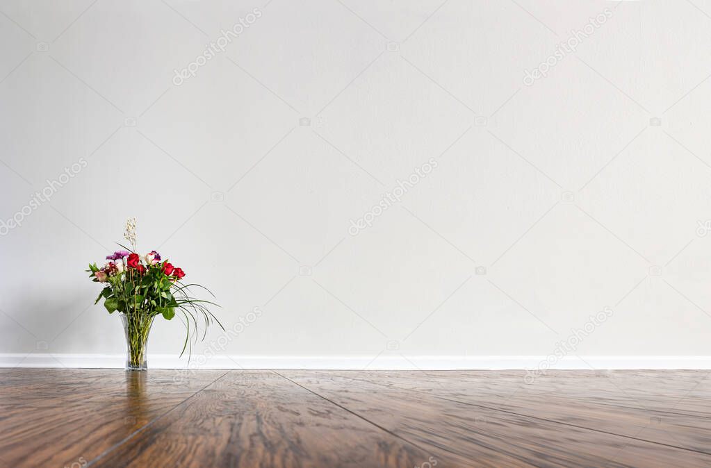Large empty room with hardwood floor, white wall and a baseboard. A vase with flower arrangement consisting of roses and long grass steams. Can be used as a background for virtual furniture or decor.