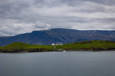 Çimlerle kaplı bir tepenin arkasındaki dağlar, Reykjavik 'e yakın olan İzlanda' nın önündeki su koyunun tam ortasından görülebilen bir ev.. 