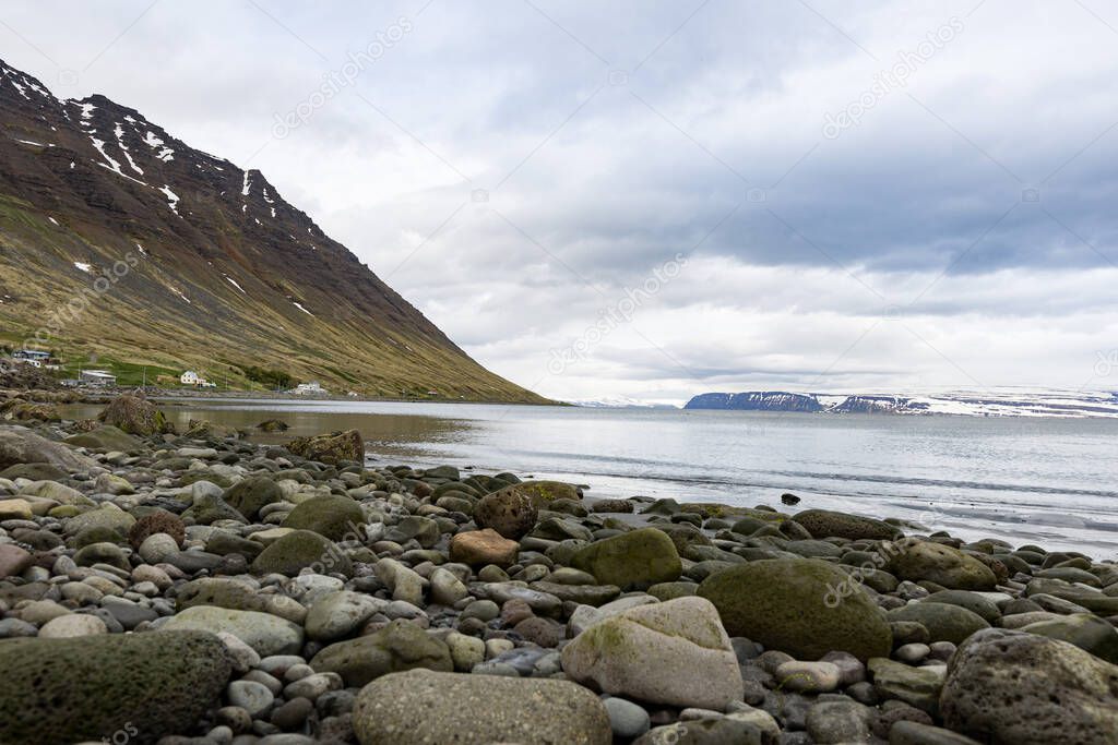 Rocks and pebbles on the shore of a fjord as seen from the village of Isafjordur, Iceland. Slope of the mountain on the left. Mountain range on the horizon.