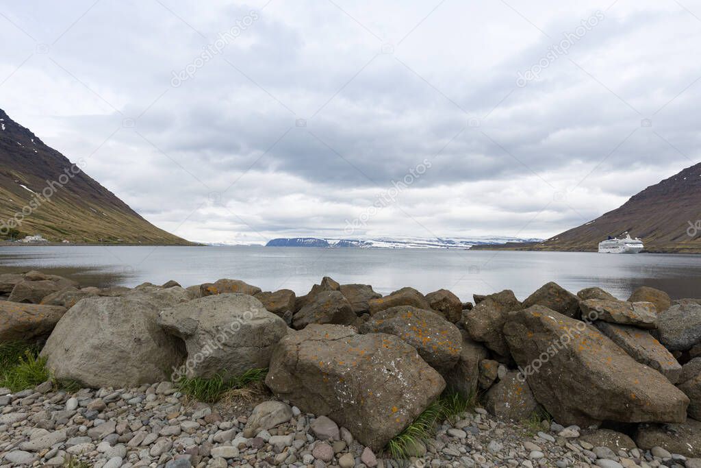 Cruise ship docked on the banks of mountains around Icelandic fjord. Pebbles and stones on the foreground, mountain ranges covered in snow on the distant horizon.
